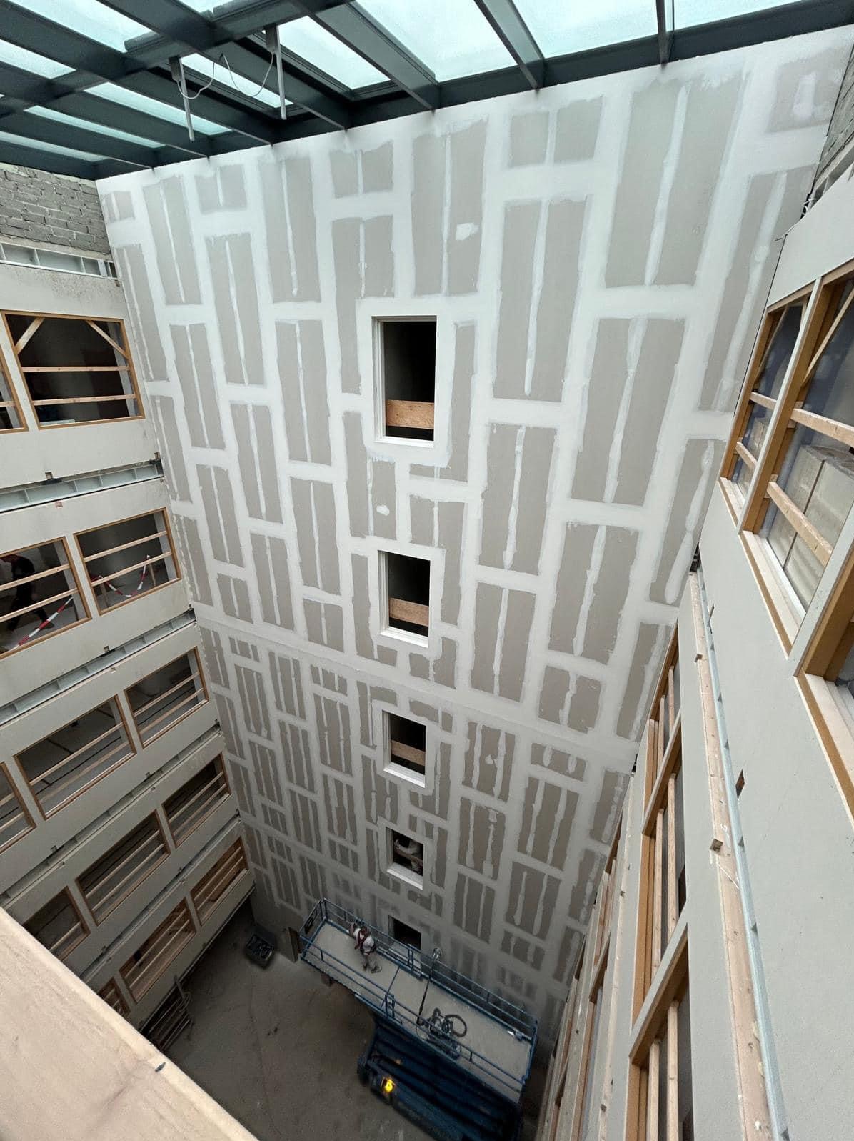 Wysoki szyb windowy w trakcie wykańczania płytami gipsowo-kartonowymi, widok od dołu z podnośnika, widoczne otwory okienne oraz szklany sufit.