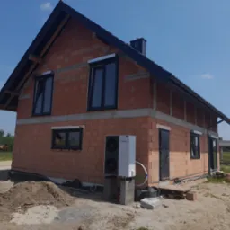 Surowy, dwukondygnacyjny dom z czerwonej cegły, z zamontowanymi oknami z roletami zewnętrznymi, czarnym dachem i kominem; na zewnątrz stoi pompa ciepła Vaillant na betonowych podstawach, widoczny...