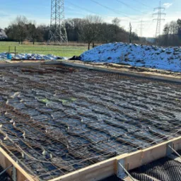 Przygotowany szalunek z siatką zbrojeniową i folią na podbudowie z izolacją kubełkową pod płytę fundamentową, w tle zasypana śniegiem skarpa ziemi i słupy wysokiego napięcia.