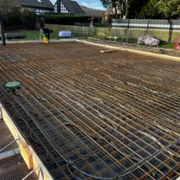 Siatka zbrojeniowa ułożona na czarnej folii w szalunku fundamentowym, widoczny pracownik w żółtych kaloszach i rura do wylewania betonu w tle.