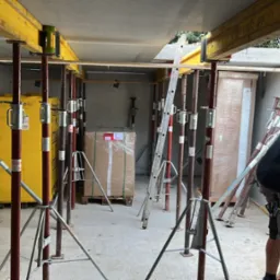 Wnętrze budynku w trakcie budowy z widocznym szalunkiem stropowym podpartym stemplami, pracownik w czarnej koszulce wchodzi po drabinie, w tle zapakowane materiały budowlane.