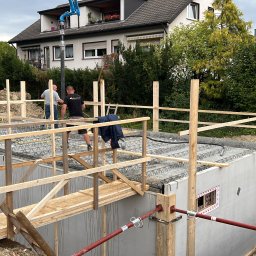 BAUFLIX - Budowa fundamentów domu jednorodzinnego: betonowe ściany, zbrojenie stropu, drewniane rusztowanie i pomost roboczy, dwóch pracowników w tle, dom w tle.