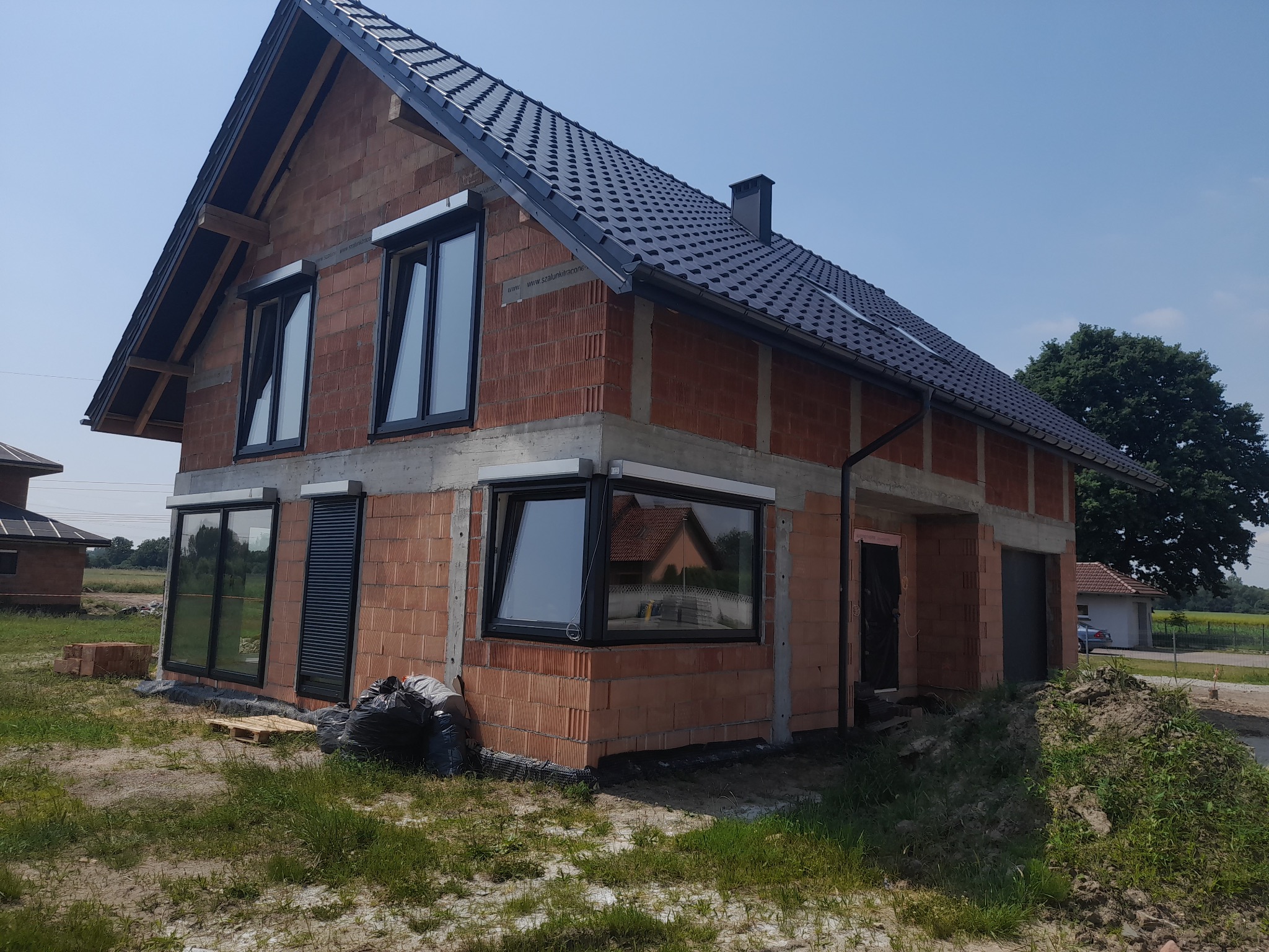 Surowy stan domu jednorodzinnego z czerwonej cegły z czarnymi oknami i grafitowym dachem, widoczne worki z odpadami budowlanymi obok palety.