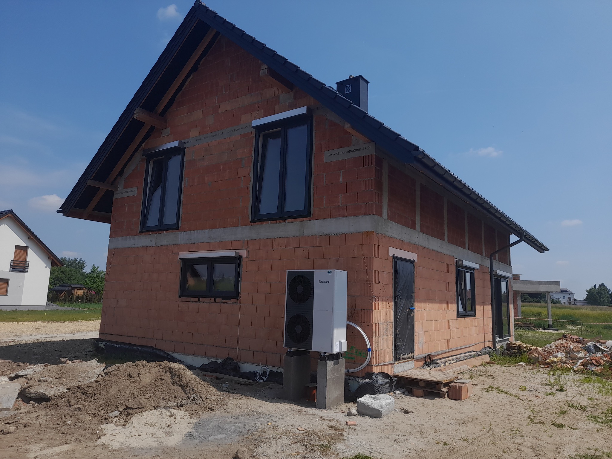Surowy, dwukondygnacyjny dom z czerwonej cegły, z zamontowanymi oknami z roletami zewnętrznymi, czarnym dachem i kominem; na zewnątrz stoi pompa ciepła Vaillant na betonowych podstawach, widoczny...