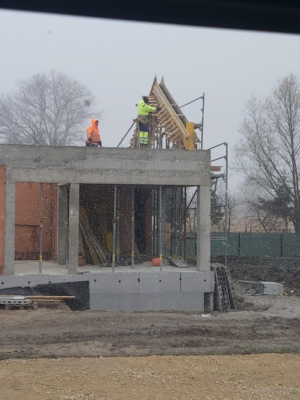 Budowa domu w trakcie zimy: dwóch robotników w odblaskowych ubraniach pracuje na szczycie surowego budynku, montując drewnianą konstrukcję dachu na tle padającego śniegu.