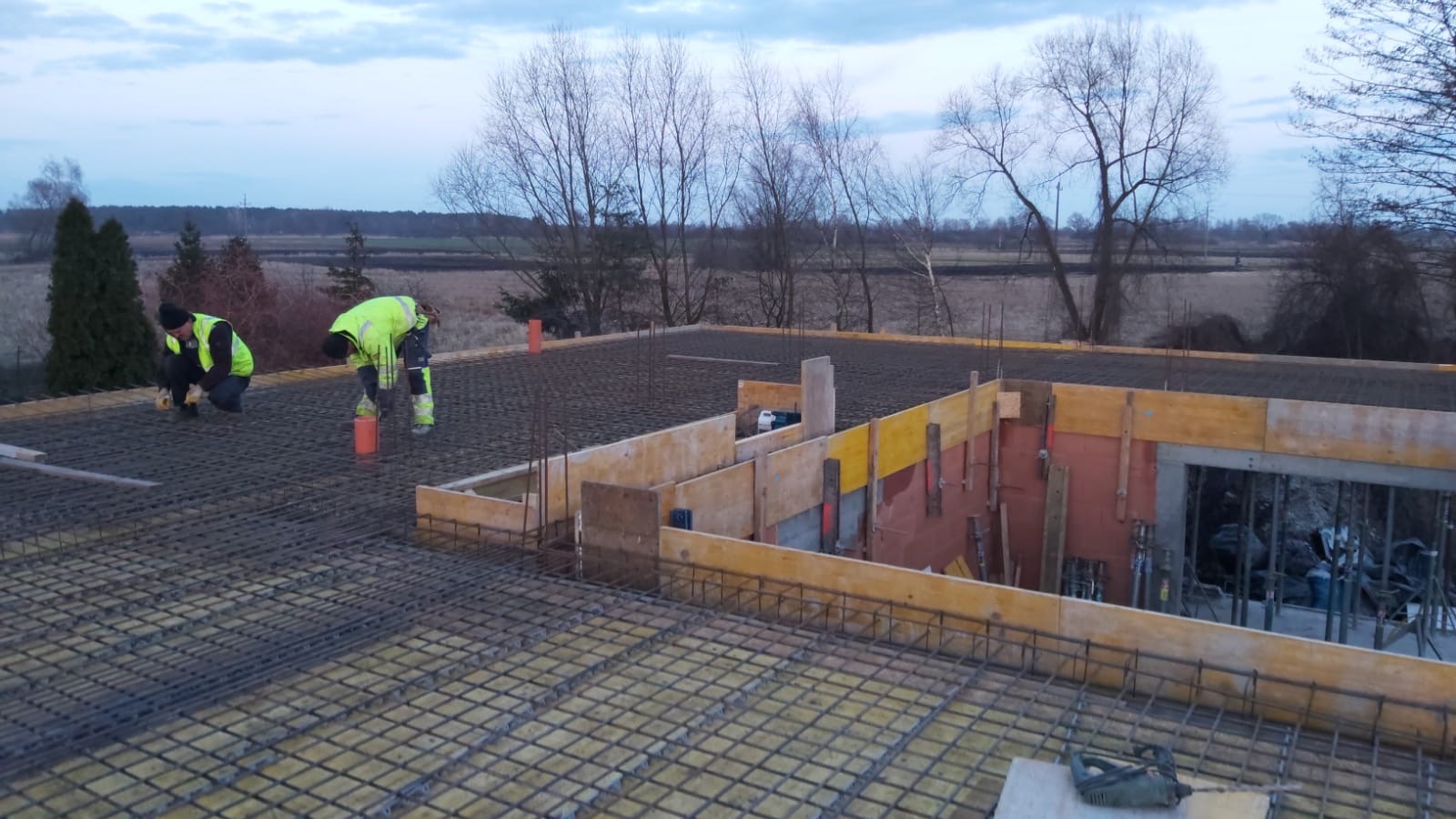 Widok z góry na plac budowy z dwoma pracownikami w odblaskowych kamizelkach układającymi siatkę zbrojeniową na przygotowanym szalunku fundamentowym, w tle widoczne drzewa i pola.