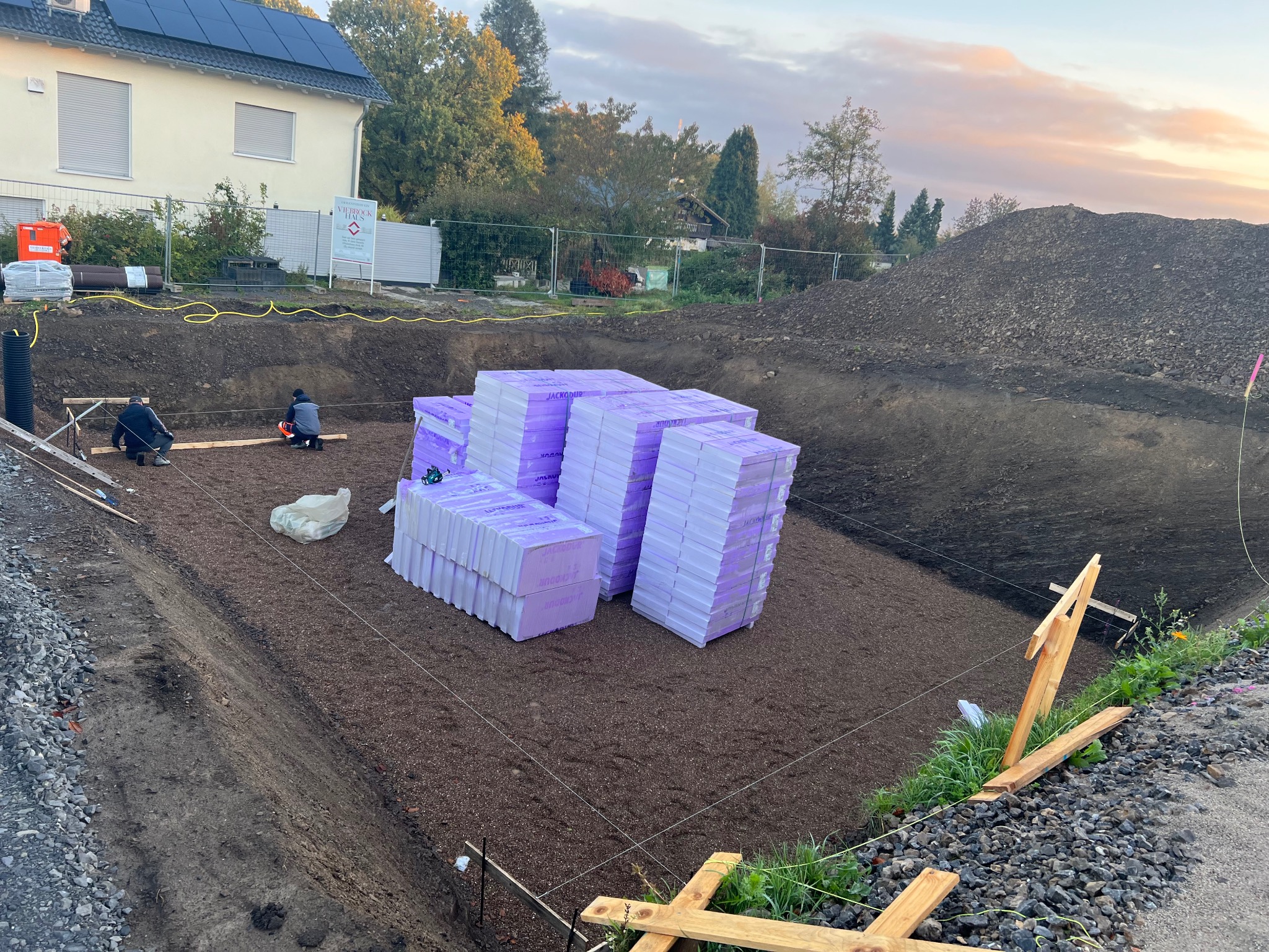 Przygotowany wykop pod budynek z ułożonym geowłókniną i podsypką, na której składowane są paczki izolacji termicznej. Dwóch pracowników przy pracy w tle, widoczny teren budowy z ogrodzeniem...