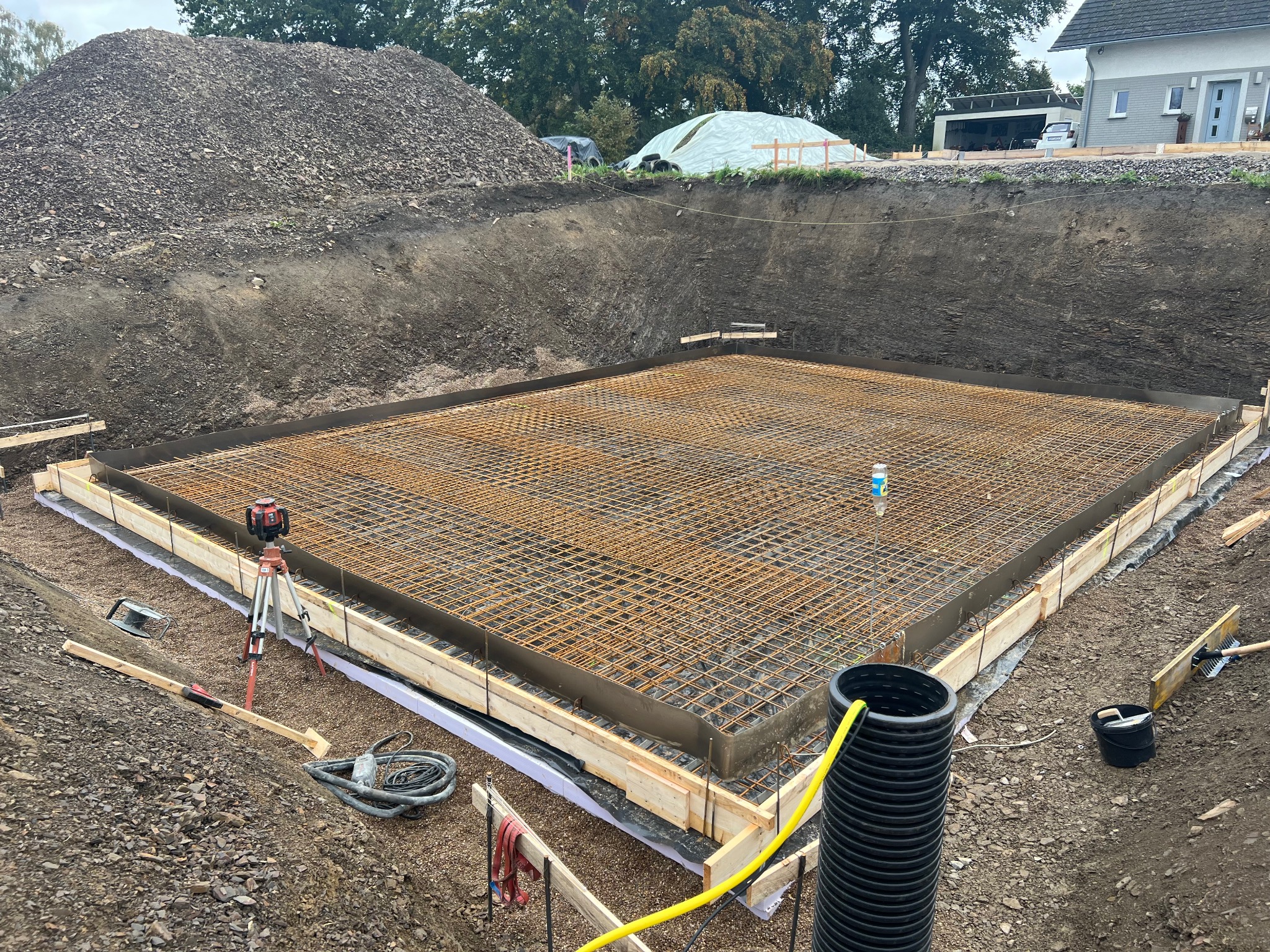 Przygotowany wykop z deskowaniem i zbrojeniem pod płytę fundamentową, widoczny niwelator laserowy na statywie, czarna rura drenażowa i duży pryzmat ziemi w tle.