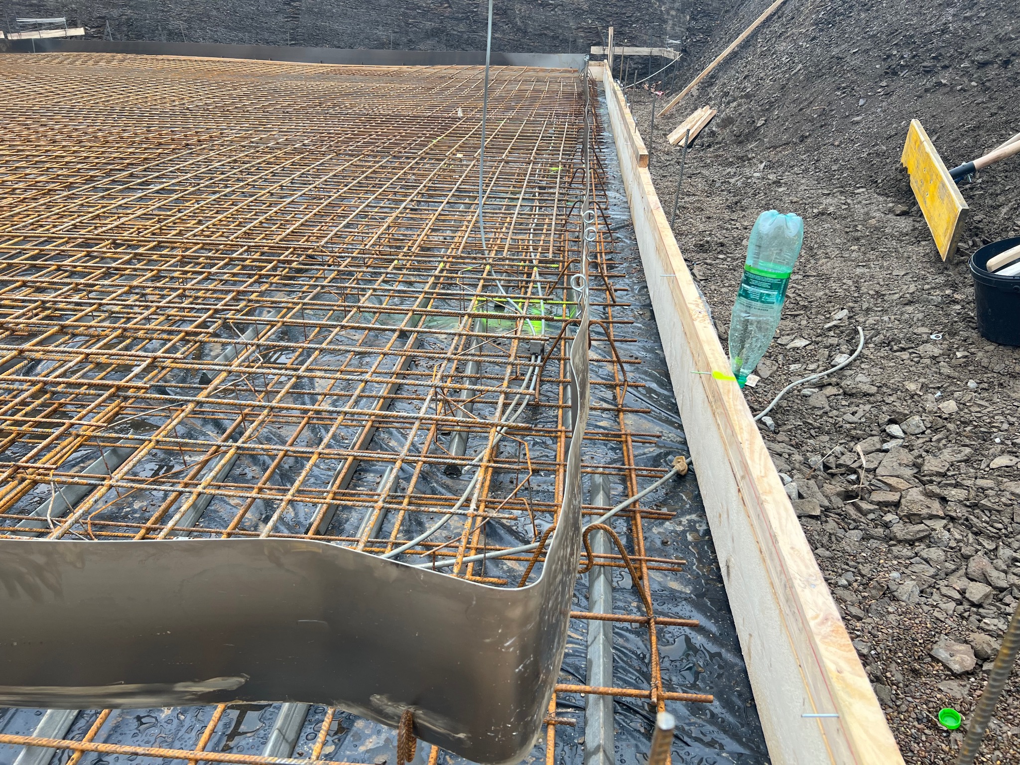 Siatka zbrojeniowa na folii w wykopie pod fundamenty, przygotowana do zalania betonem, widoczne szalunki i butelka wody obok łopaty.