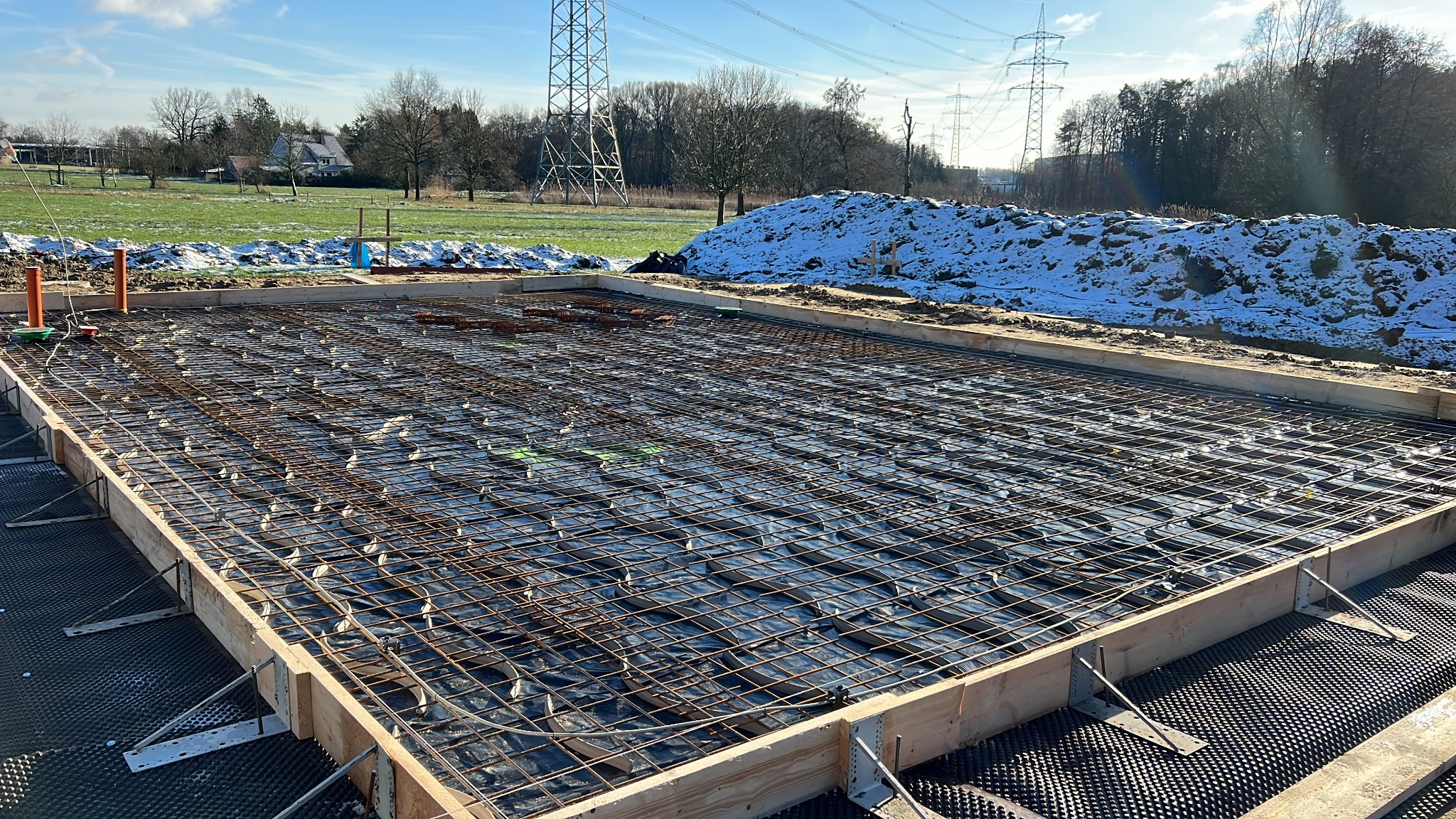 Przygotowany szalunek z siatką zbrojeniową i folią na podbudowie z izolacją kubełkową pod płytę fundamentową, w tle zasypana śniegiem skarpa ziemi i słupy wysokiego napięcia.