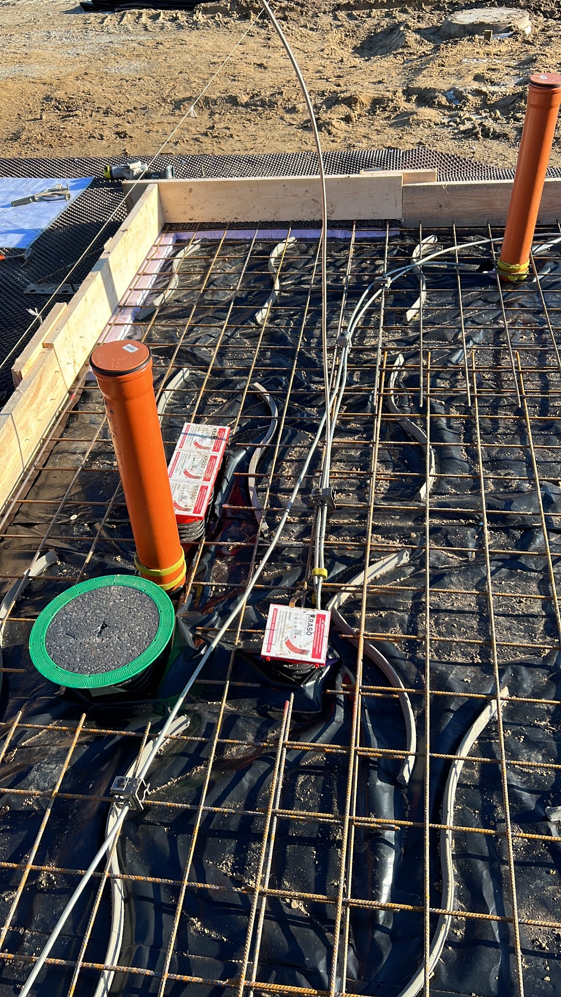 Zbrojenie fundamentu z ułożonymi rurami kanalizacyjnymi i przewodami elektrycznymi na czarnej folii, widoczne drewniane szalunki i grunt w tle.