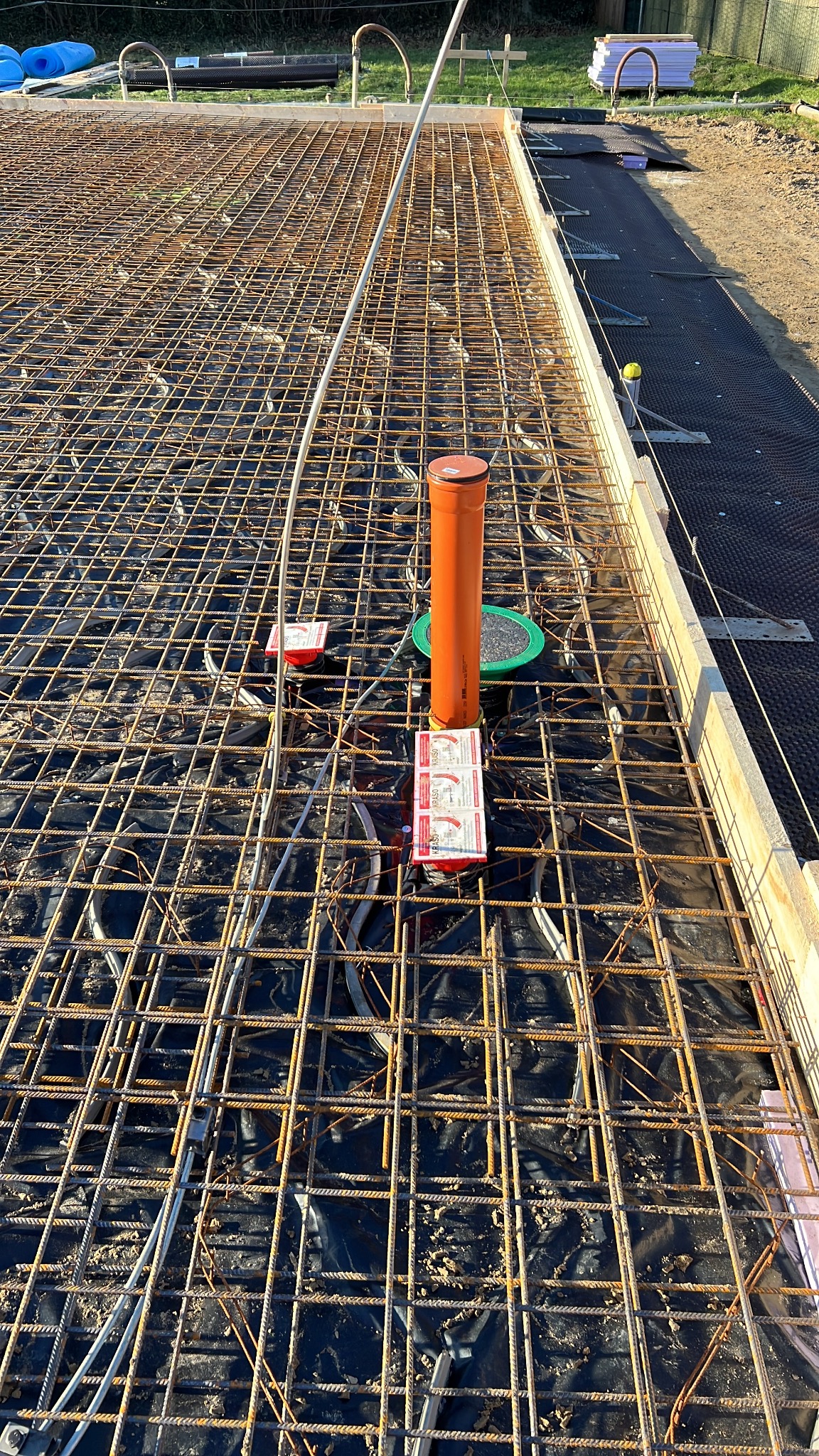 Siatka zbrojeniowa przygotowana pod wylewkę fundamentową, z widoczną rurą kanalizacyjną i elementami dystansowymi Krasso, ułożona na czarnej folii hydroizolacyjnej.