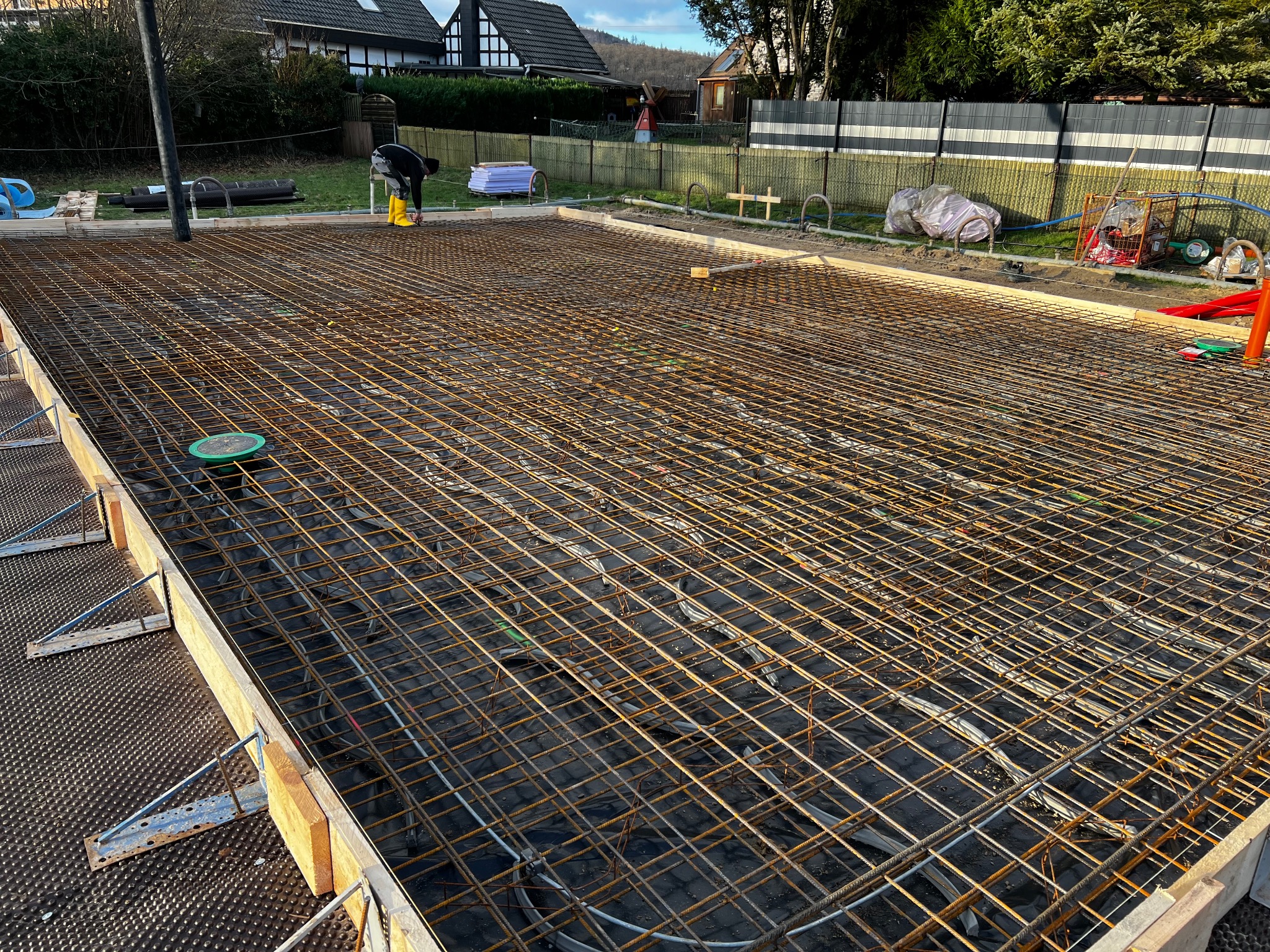 Siatka zbrojeniowa ułożona na czarnej folii w szalunku fundamentowym, widoczny pracownik w żółtych kaloszach i rura do wylewania betonu w tle.