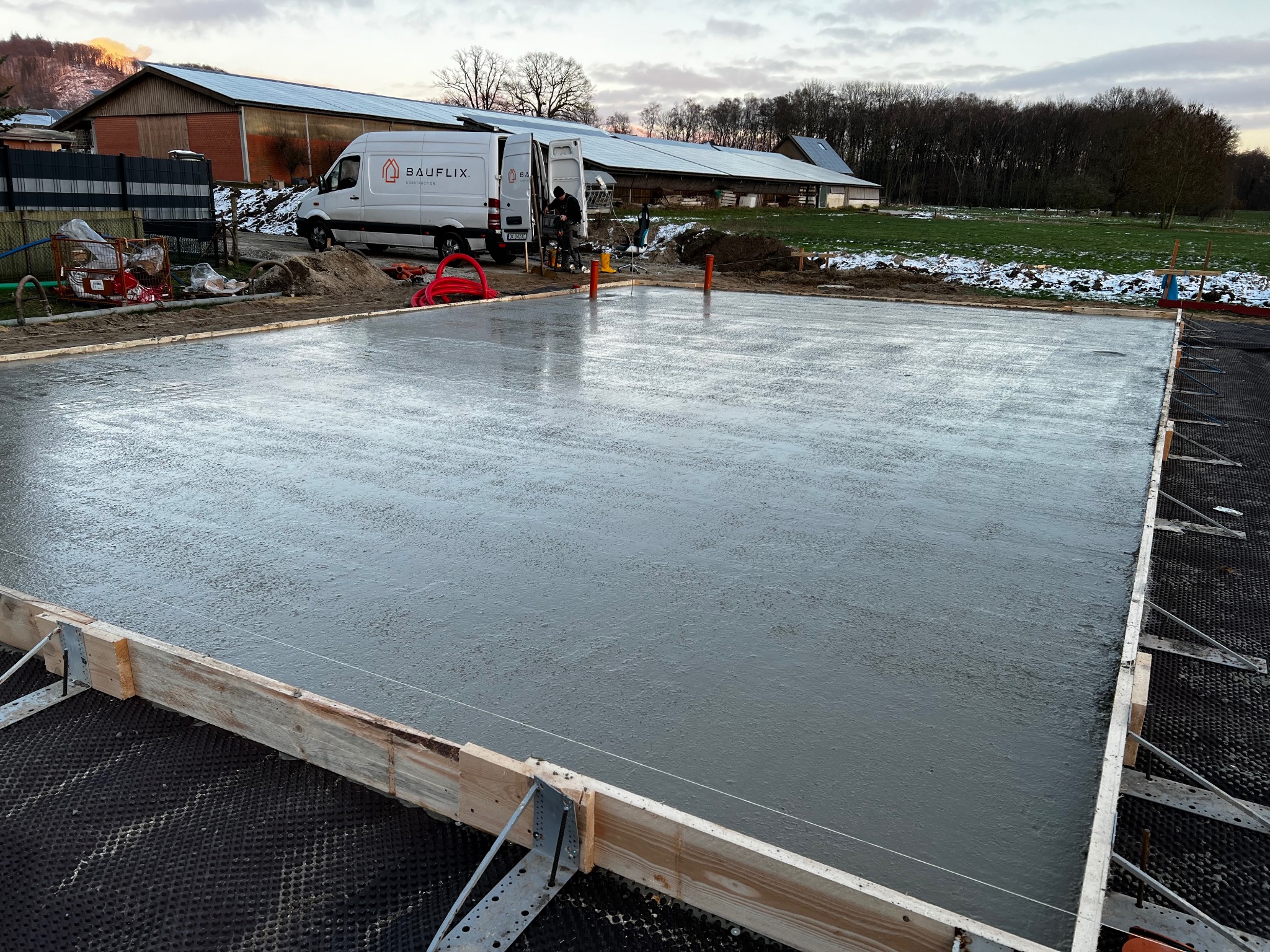 Świeżo wylana, gładka betonowa płyta fundamentowa w drewnianym szalunku, widoczny firmowy van BÄUFLIX w tle na tle pochmurnego nieba i częściowo ośnieżonego terenu.