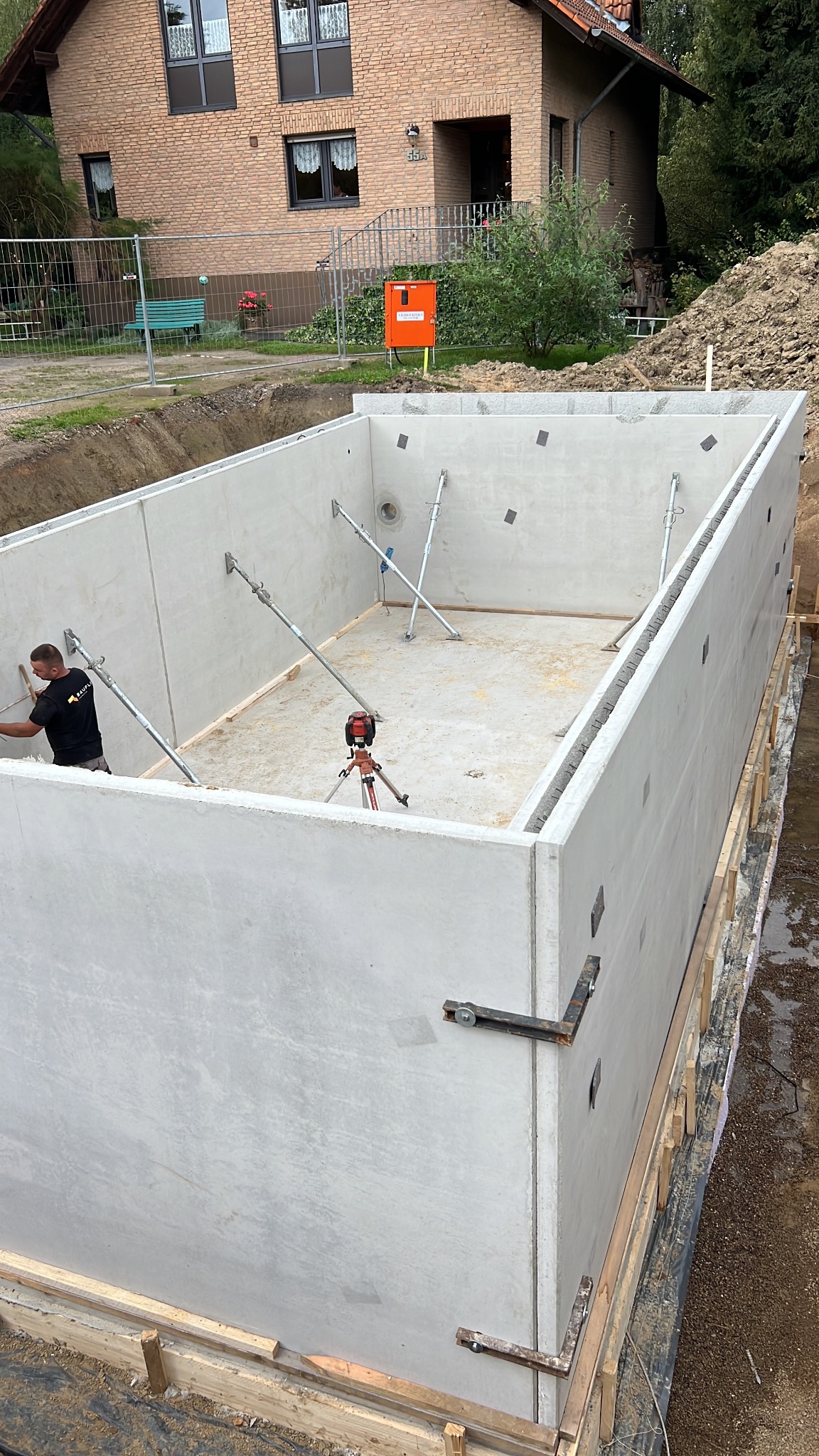 Wykopany fundament z betonowymi ścianami szalunkowymi wzmocnionymi metalowymi rozpórkami, widoczny pracownik w masce i niwelator laserowy na statywie.