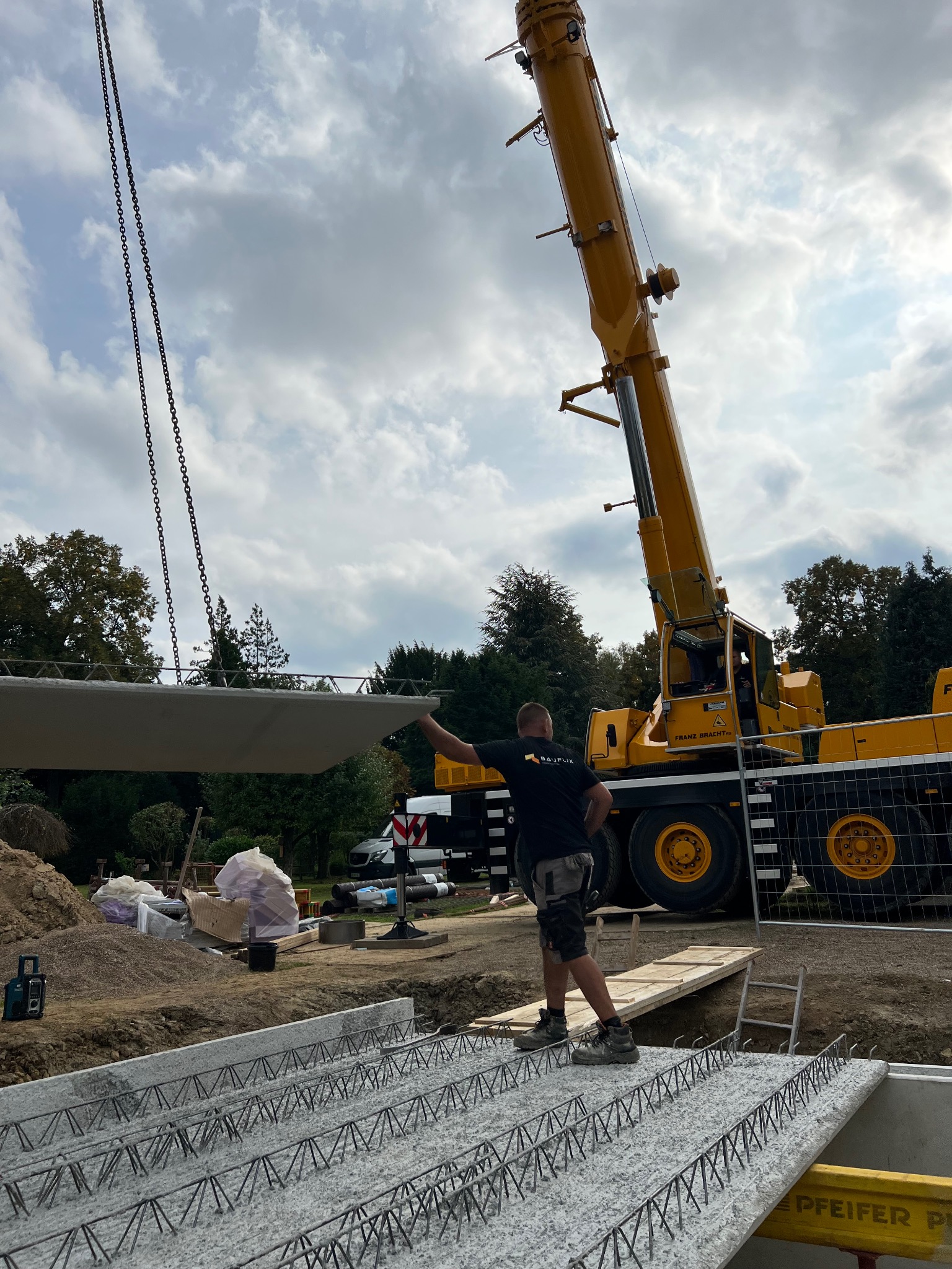 Żuraw budowlany opuszcza prefabrykowany element stropu żelbetowego na przygotowane zbrojenie fundamentu; widoczny pracownik kierujący operacją, teren budowy z zielenią w tle.