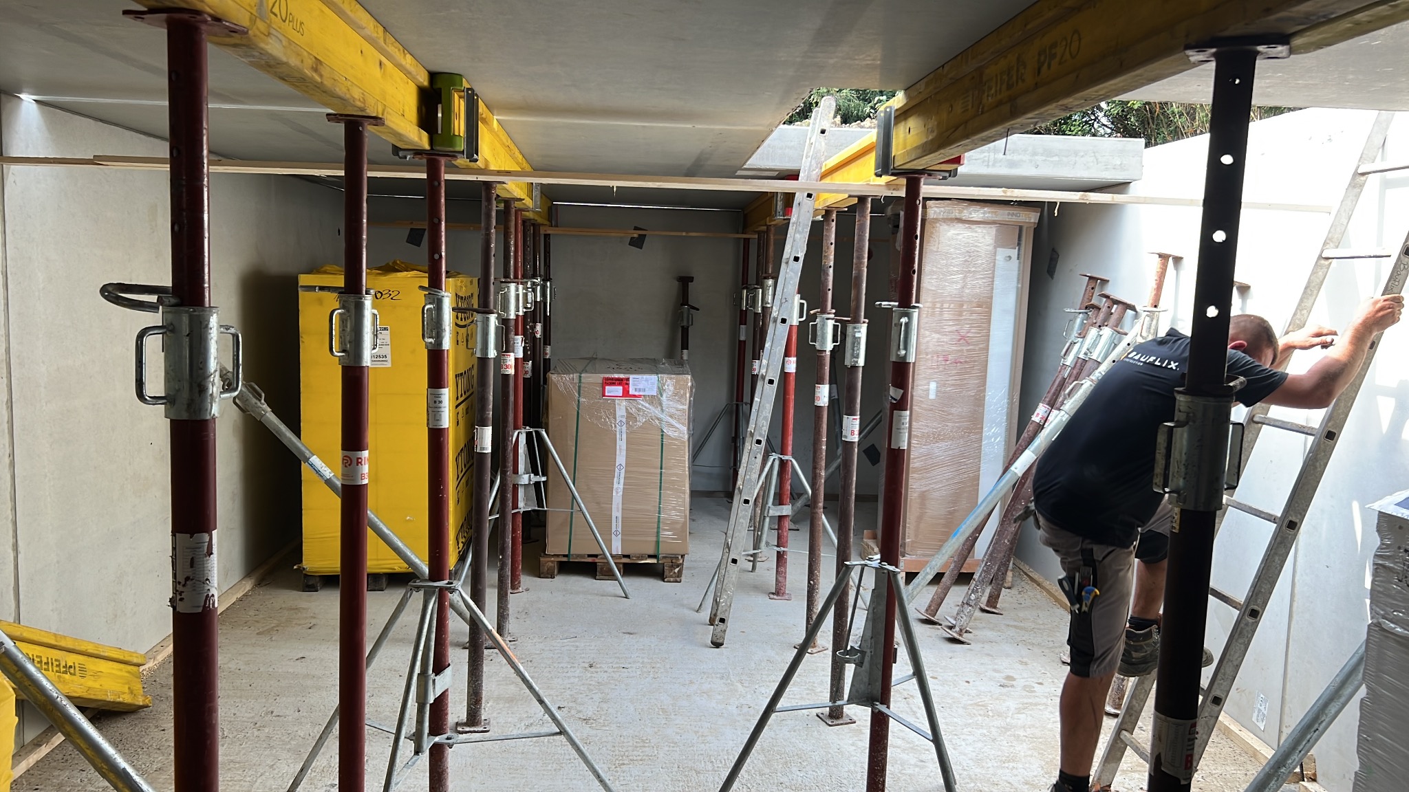 Wnętrze budynku w trakcie budowy z widocznym szalunkiem stropowym podpartym stemplami, pracownik w czarnej koszulce wchodzi po drabinie, w tle zapakowane materiały budowlane.