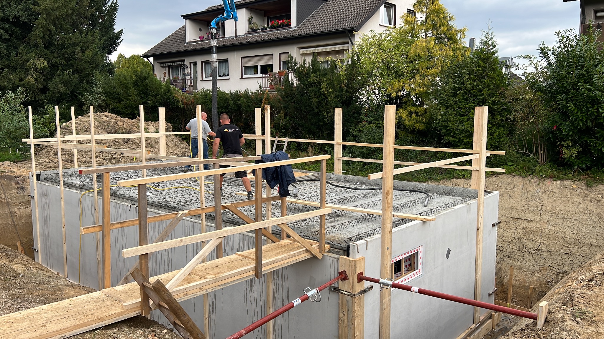 Budowa fundamentów domu jednorodzinnego: betonowe ściany, zbrojenie stropu, drewniane rusztowanie i pomost roboczy, dwóch pracowników w tle, dom w tle.