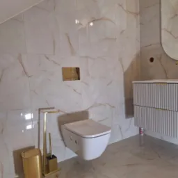 Nowoczesna łazienka z beżowo-złotymi płytkami imitującymi marmur, podwieszana toaleta, złote akcesoria łazienkowe i umywalka z szafką w stylu glamour.