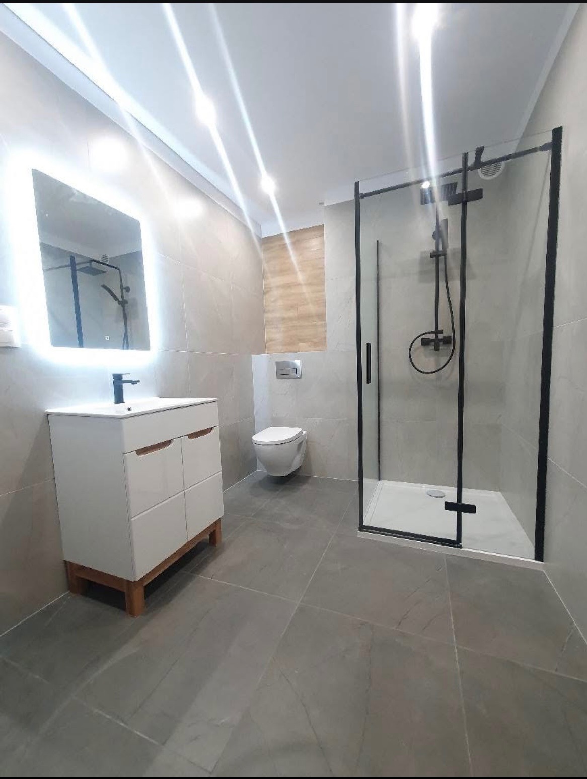 Nowoczesna łazienka z szarymi płytkami, białą szafką pod umywalkę z drewnianymi nóżkami i czarną kabiną prysznicową z deszczownicą.