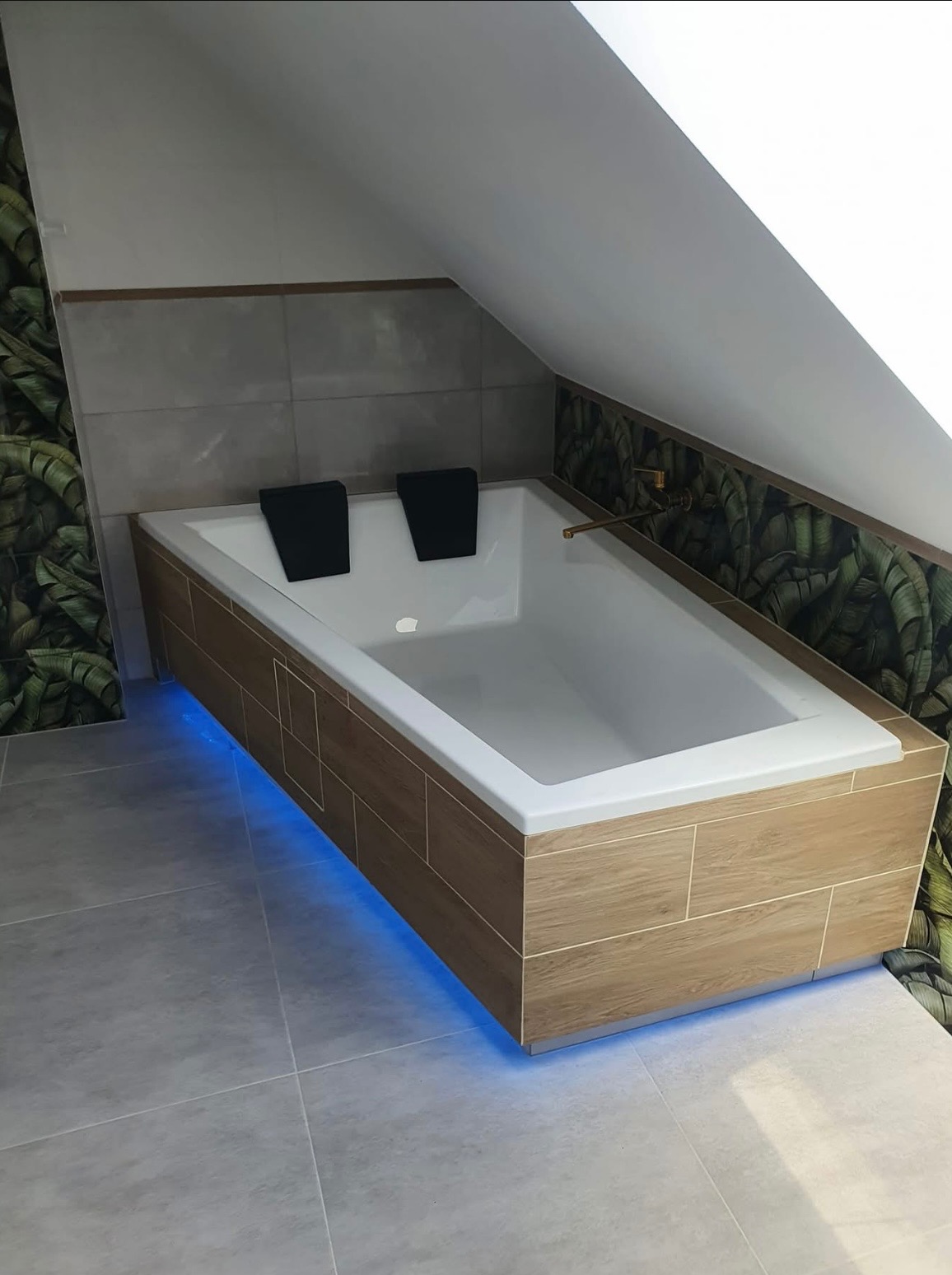 Nowoczesna wanna w obudowie z płytek imitujących drewno, podświetlona niebieskim światłem LED, z czarnymi zagłówkami, w łazience z motywem tropikalnych liści na ścianie.