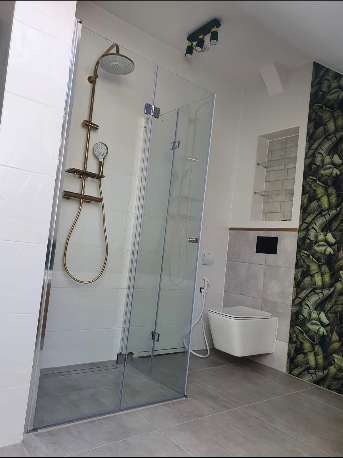 Nowoczesna łazienka z kabiną prysznicową ze złotym zestawem natryskowym, wisząca toaleta i ściana z motywem liści bananowca.