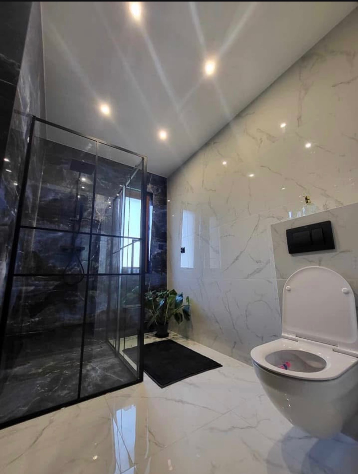 Nowoczesna łazienka z kabiną prysznicową typu walk-in z czarnymi ramami, marmurowe płytki na ścianach i podłodze, wisząca toaleta z czarnym panelem sterującym spłuczką, roślina doniczkowa i czarny...