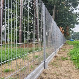 Marbruk - Długa, metalowa siatka ogrodzeniowa z betonową podmurówką, widoczna z boku na tle zieleni i drzew.