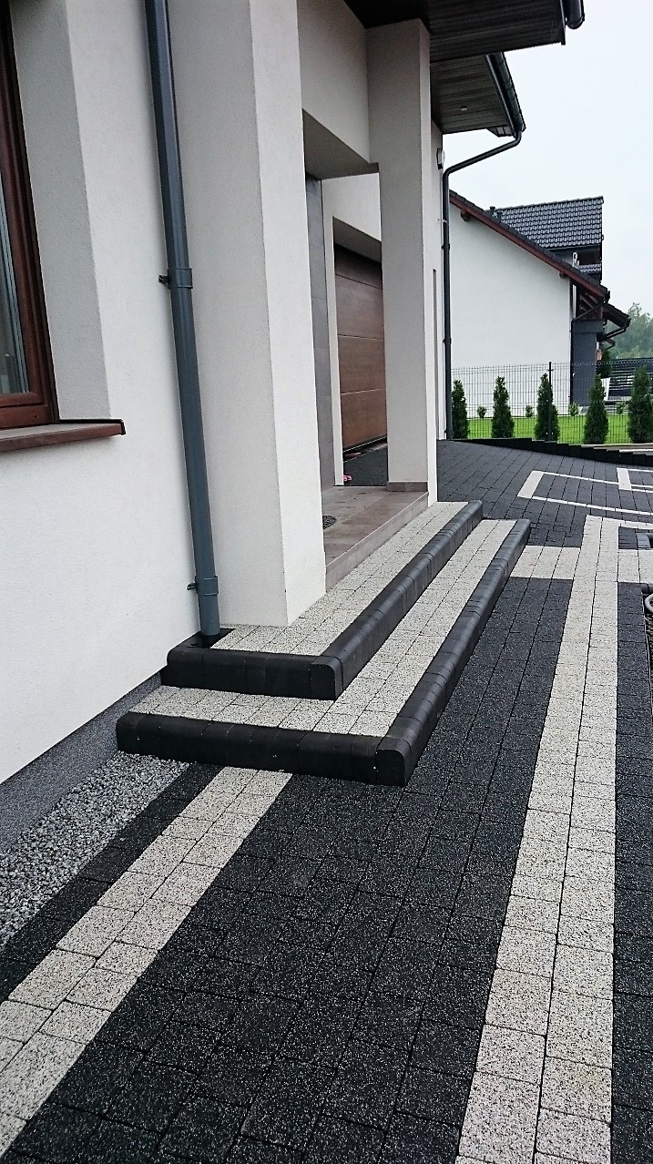 Nowoczesne wejście do domu z kostki brukowej w kolorach szarości, schody wykończone czarnym obrzeżem, widok na biały budynek i starannie utrzymany ogród.