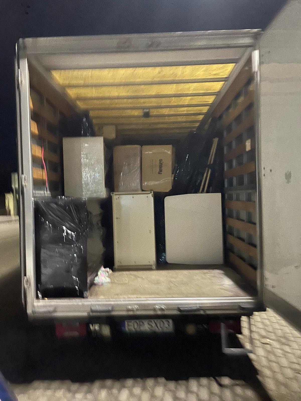 Wnętrze furgonetki załadowane meblami i kartonami, niektóre owinięte folią, widok z tyłu pojazdu, tablica rejestracyjna EOP SX03.