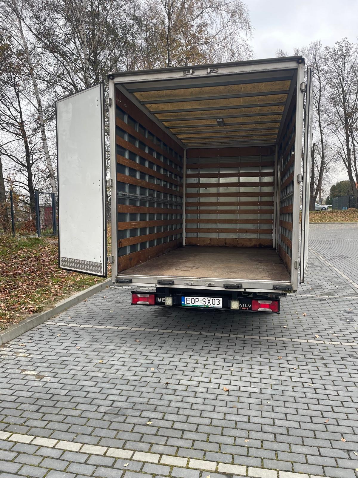 Pusta przestrzeń ładunkowa białego samochodu dostawczego z otwartymi tylnymi drzwiami, widok z tyłu na parkingu z kostki brukowej w pochmurny dzień.