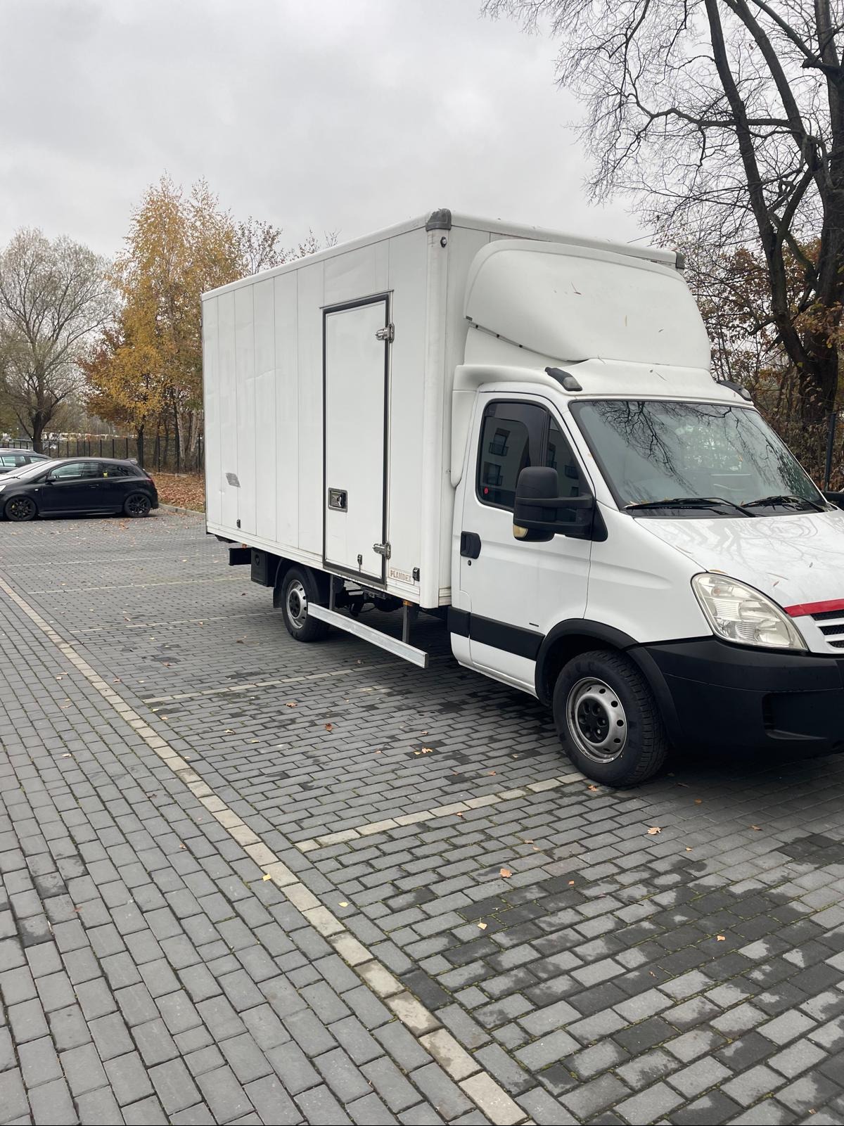 Biały furgon dostawczy zaparkowany na brukowanym parkingu w pochmurny dzień, widoczne jesienne liście na ziemi.