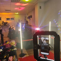 Paweł Łukasik - Fotobudka w akcji: smartfon w obudowie z oświetleniem LED, w tle para gości i dekoracje balonowe, światła dyskotekowe na suficie, rekwizyty na stole.