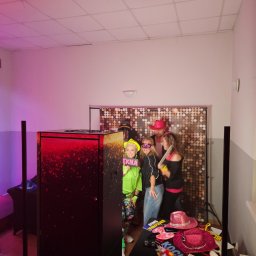 Paweł Łukasik - Grupa osób pozuje do zdjęcia w fotobudce z błyszczącym tłem i rekwizytami, takimi jak kapelusze i tabliczki z napisami. Widoczna fotobudka z czerwonym oświetleniem.