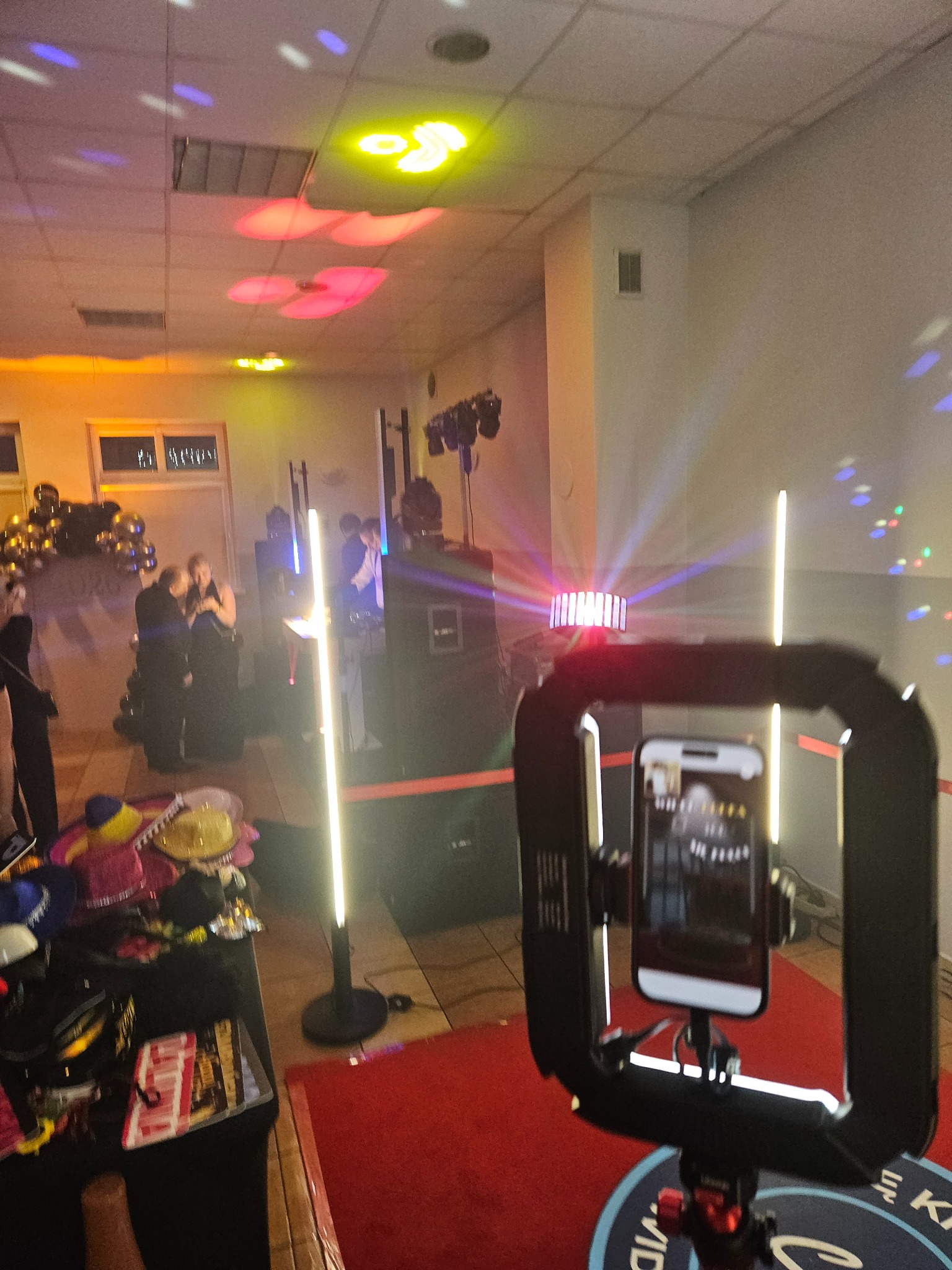 Fotobudka w akcji: smartfon w obudowie z oświetleniem LED, w tle para gości i dekoracje balonowe, światła dyskotekowe na suficie, rekwizyty na stole.