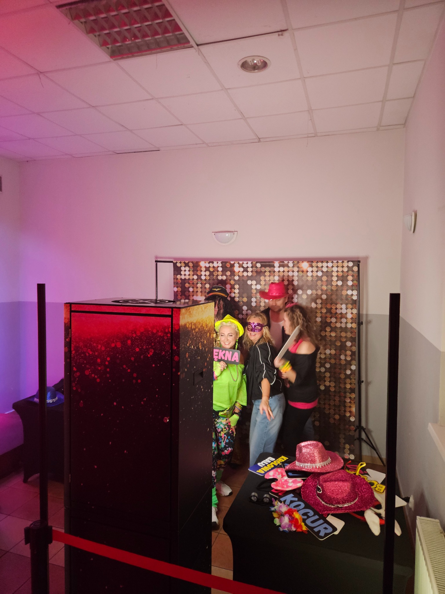 Grupa osób pozuje do zdjęcia w fotobudce z błyszczącym tłem i rekwizytami, takimi jak kapelusze i tabliczki z napisami. Widoczna fotobudka z czerwonym oświetleniem.