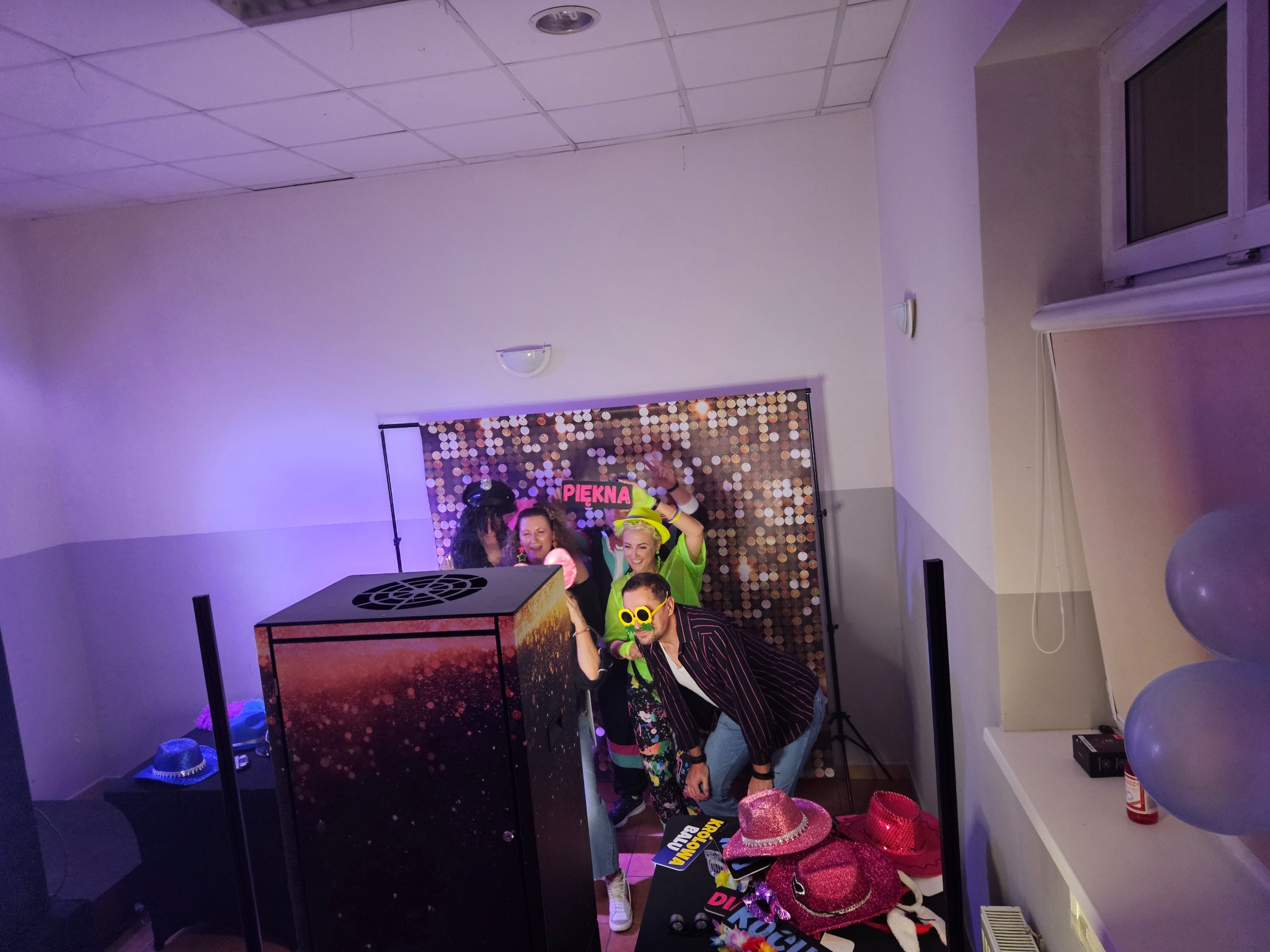 Grupa osób w przebraniach pozuje do zdjęcia przy fotobudce z tłem w cekiny. Na pierwszym planie rekwizyty: kapelusze i tabliczki z napisami. Fioletowe oświetlenie.