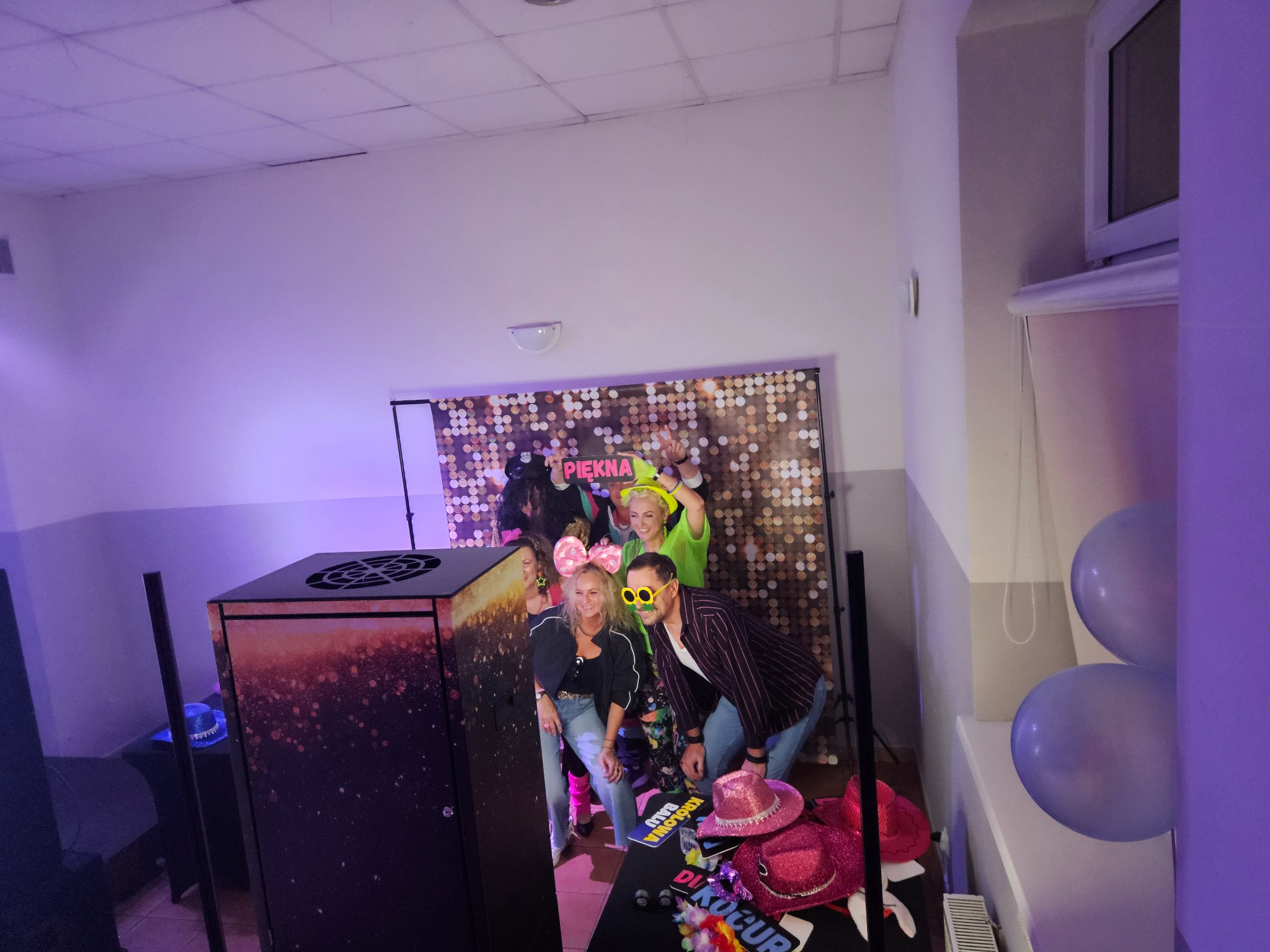Grupa osób w neonowych przebraniach pozuje przy fotobudce z błyszczącym tłem, rekwizytami i balonami, tworząc radosną atmosferę imprezy.