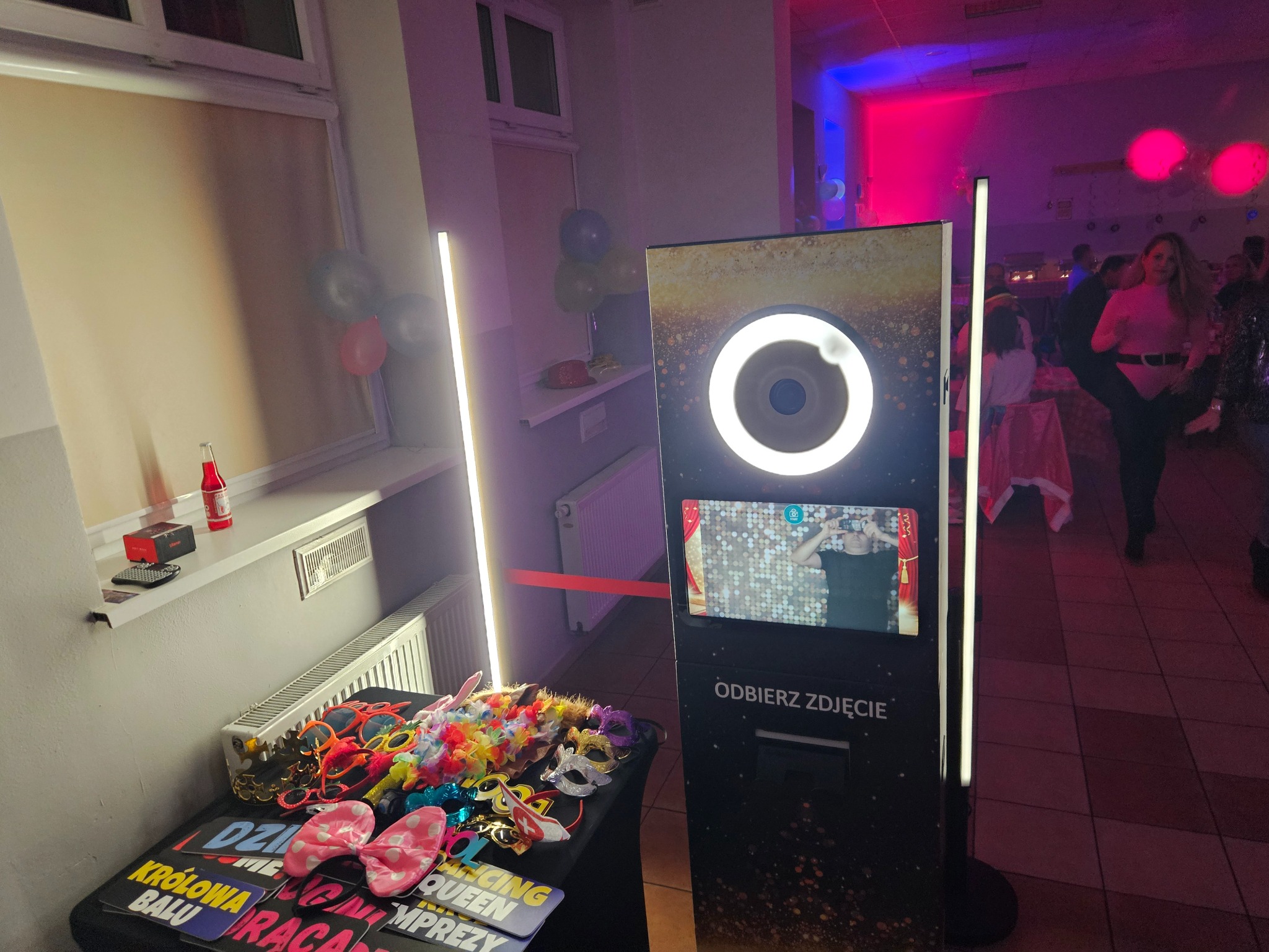 Fotobudka z rekwizytami (maski, okulary, tabliczki z napisami) na stole, oświetlona lampami LED, z widocznym ekranem i napisem 'Odbierz zdjęcie'.