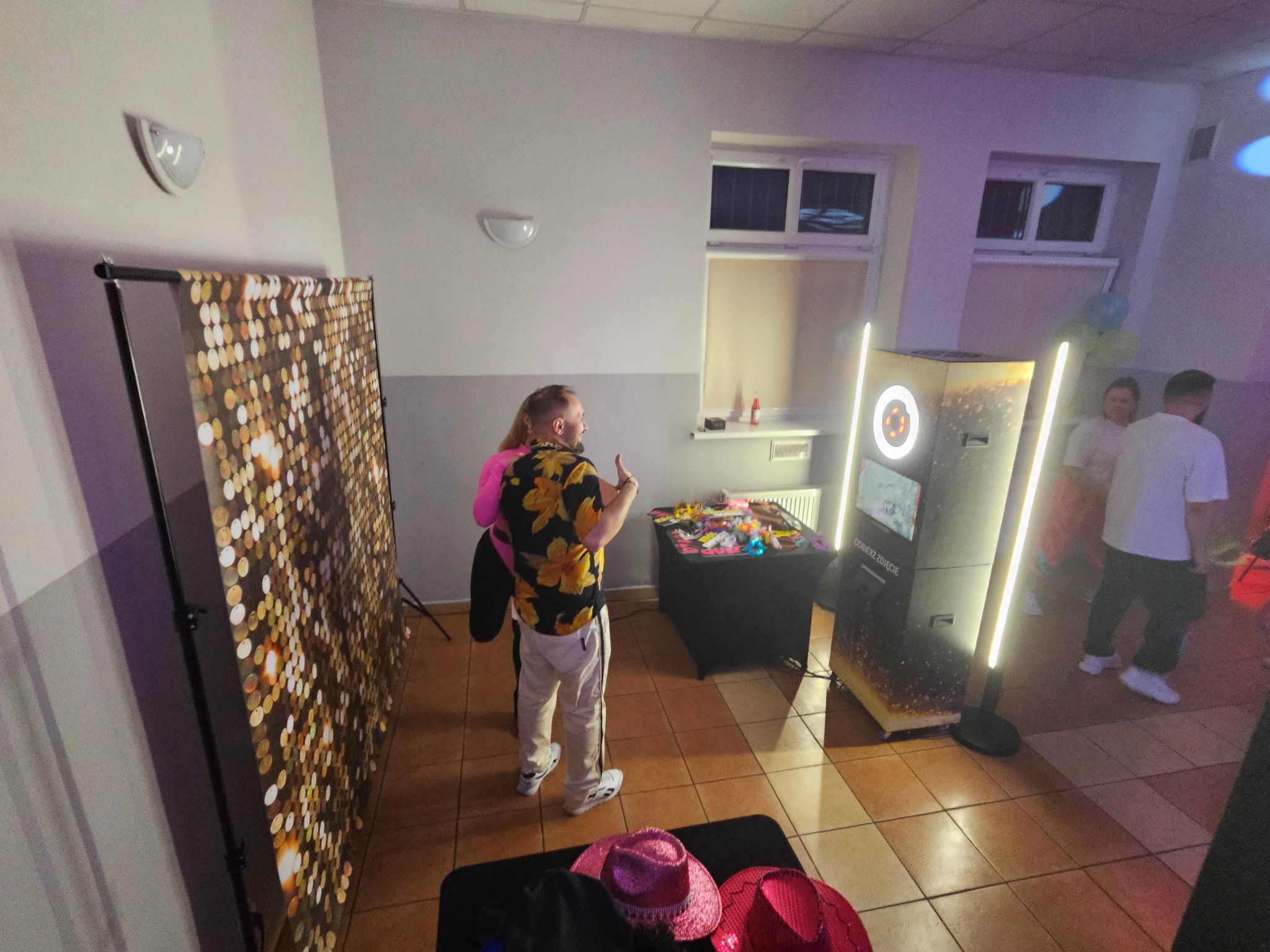 Uczestnicy imprezy pozują przy fotobudce z błyszczącym tłem i rekwizytami, w tle widoczni inni goście. Oświetlenie imprezowe tworzy klimat zabawy.