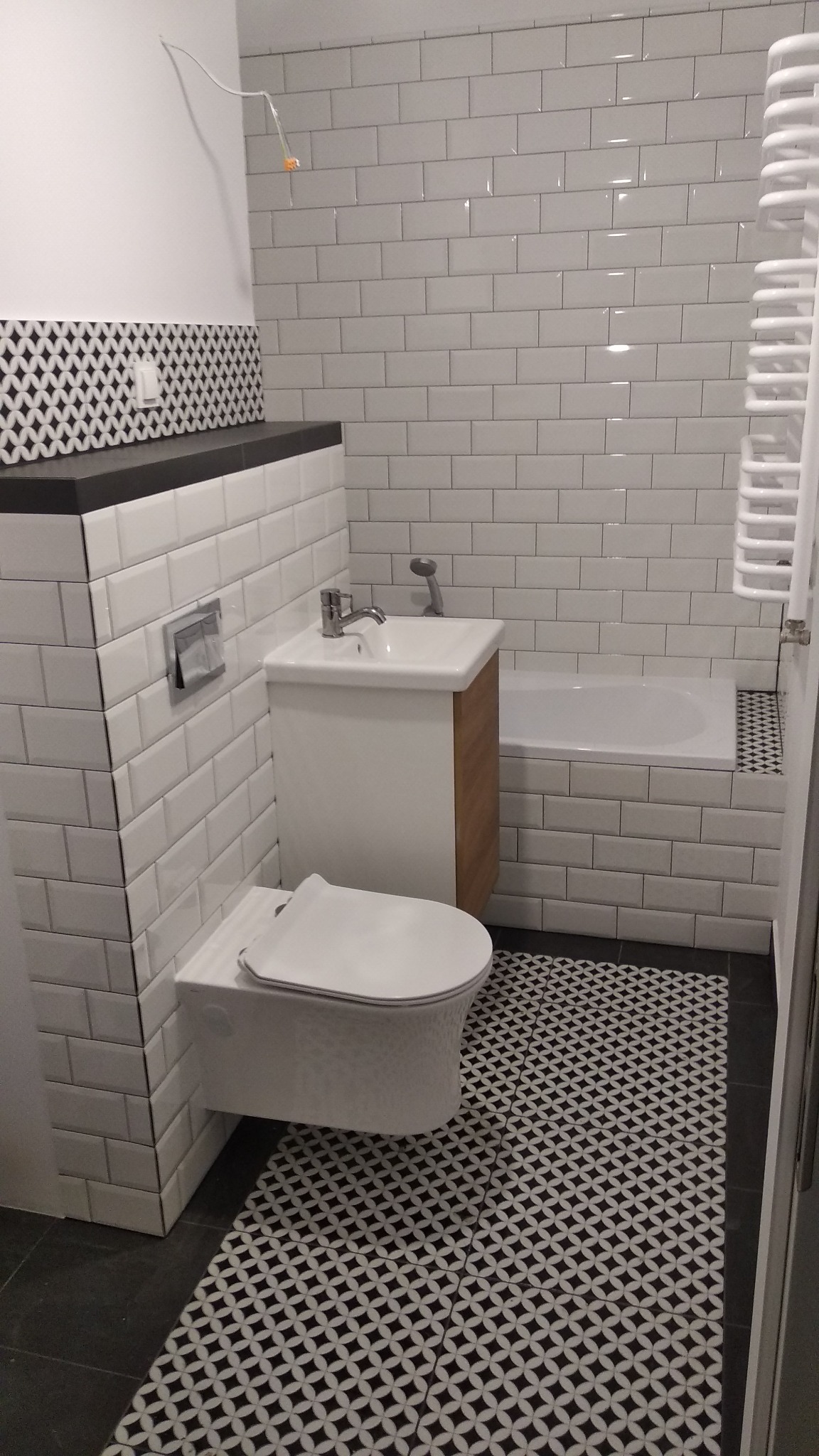 Nowoczesna łazienka z białymi płytkami typu metro na ścianach, umywalką z szafką z drewnianymi elementami, podwieszaną toaletą i podłogą w czarno-biały wzór geometryczny.