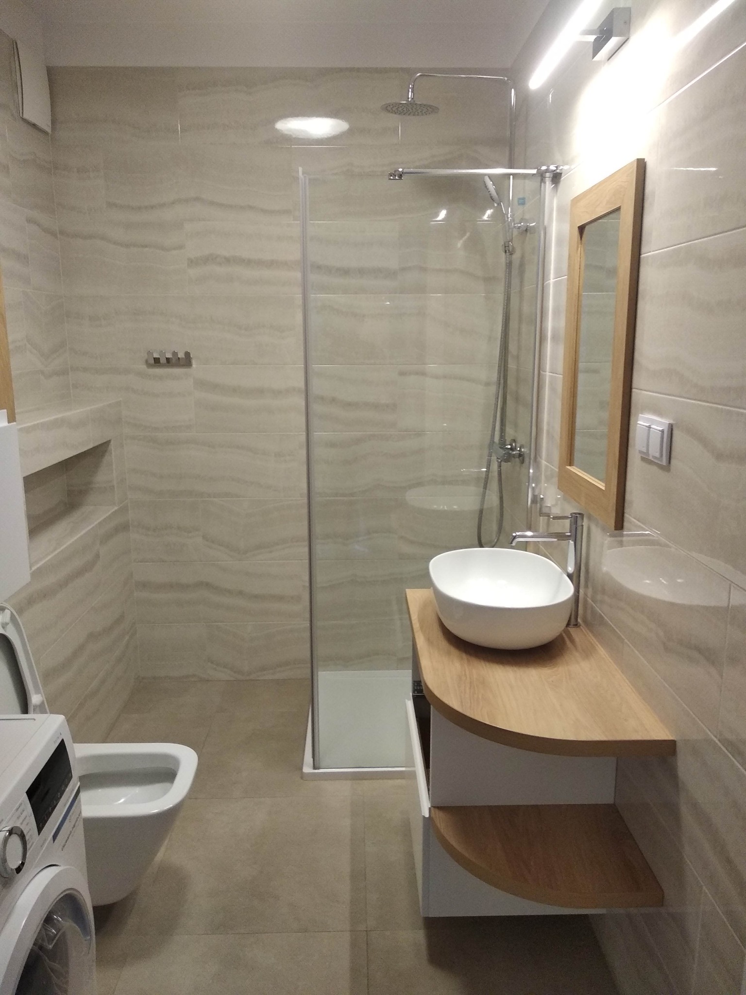 Jasna łazienka z kabiną prysznicową, umywalką nablatową na drewnianym blacie, toaletą podwieszaną i pralką z przodu.