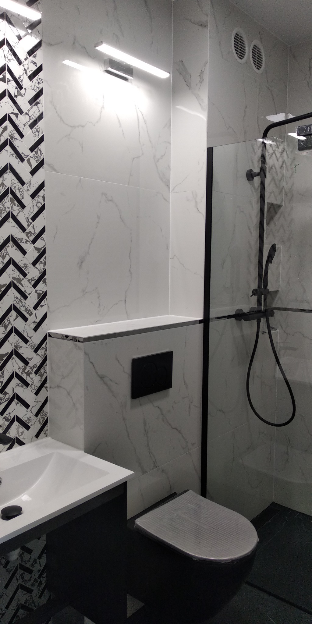 Nowoczesna łazienka z czarno-białymi płytkami w jodełkę, marmurowymi płytami ściennymi, czarną armaturą i podwieszaną miską WC z zabezpieczeniem na desce.