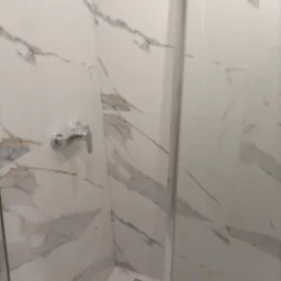 Kabina prysznicowa wykończona białymi płytkami imitującymi marmur, z widoczną chromowaną baterią i odpływem liniowym w podłodze.