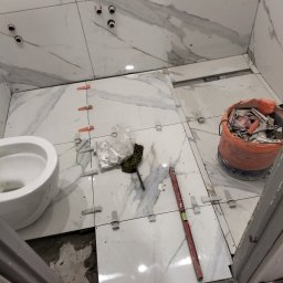 Ar-bud - Białe, marmurkowe płytki podłogowe w trakcie układania w łazience, widoczne poziomica, krzyżyki dystansowe i wiadro z odpadami, przygotowane otwory na rury.