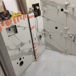 Ar-bud - Białe płytki imitujące marmur, ułożone na ścianie łazienki w trakcie prac wykończeniowych; widoczne plastikowe krzyżyki dystansowe, otwory na instalacje i fragment ściany z instalacją podtynkową.