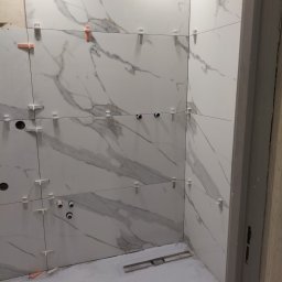 Ar-bud - Ściany łazienki w trakcie kładzenia płytek imitujących marmur, z widocznymi plastikowymi krzyżykami dystansowymi i poziomnicą na podłodze.