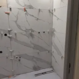 Ściany łazienki w trakcie kładzenia płytek imitujących marmur, z widocznymi plastikowymi krzyżykami dystansowymi i poziomnicą na podłodze.