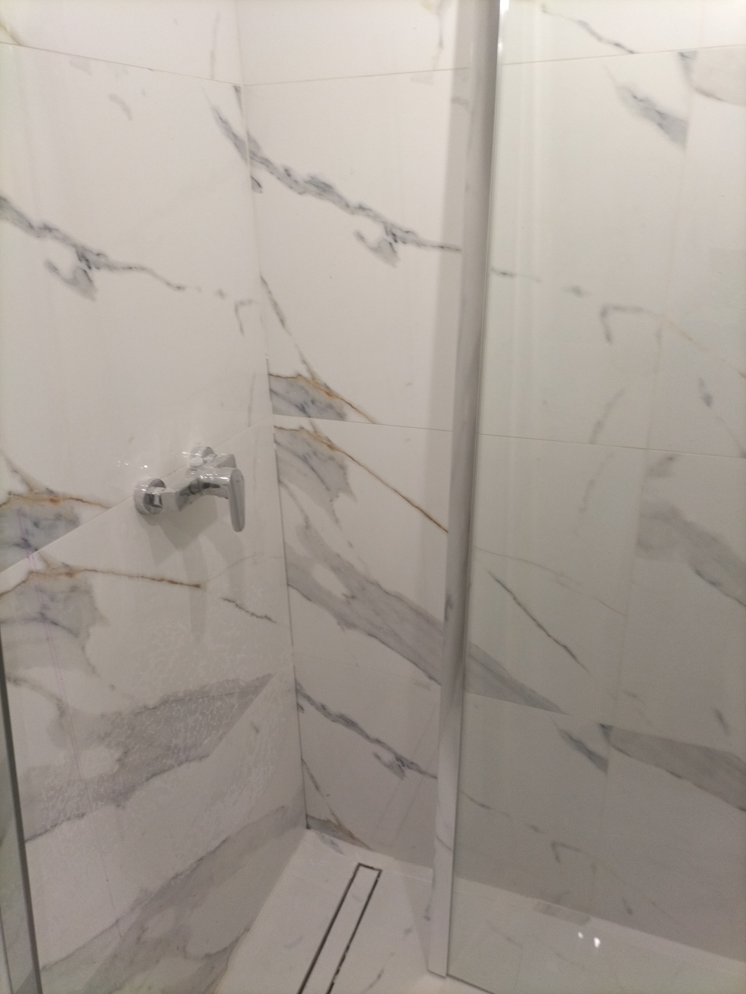 Kabina prysznicowa wykończona białymi płytkami imitującymi marmur, z widoczną chromowaną baterią i odpływem liniowym w podłodze.