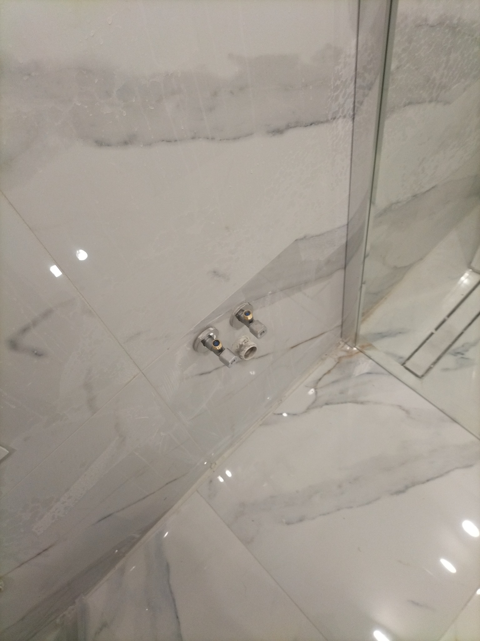 Narożnik kabiny prysznicowej wyłożony marmurkową glazurą, widoczne zawory i odpływ liniowy, silikonowanie łączeń.